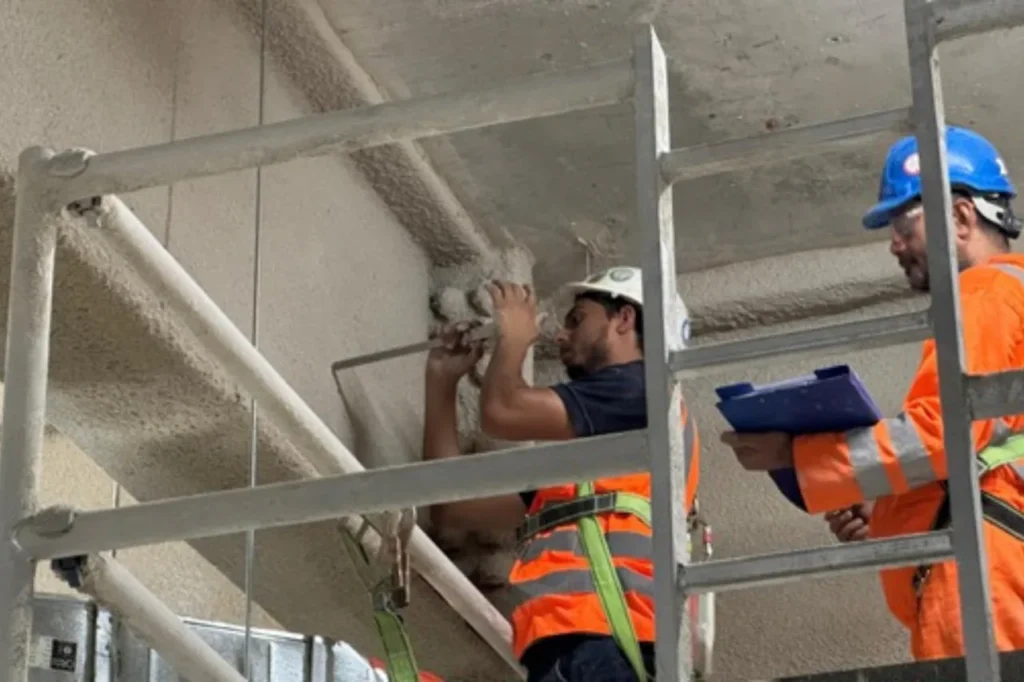 Third-party inspectors checking spray fireproofing on structural steel from a scaffold
